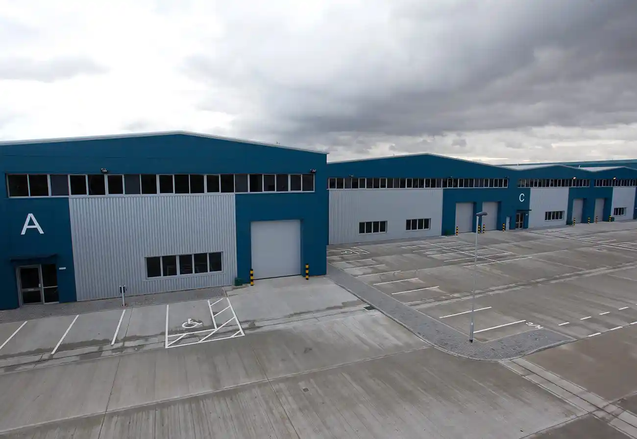 Modern industrial warehouse units with blue and grey facades, marked with letters "A" and "C," featuring large roller doors, multiple windows, and ample empty parking spaces under a cloudy sky.