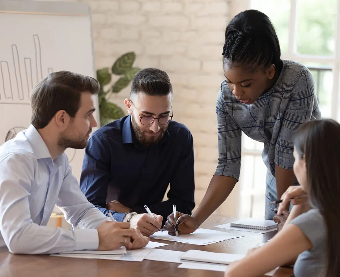 Diverse team of four professionals collaborating around a table in a bright office, discussing documents and taking notes, illustrating Cudd Bentley Consulting's collaborative and inclusive approach to sustainable consulting solutions.