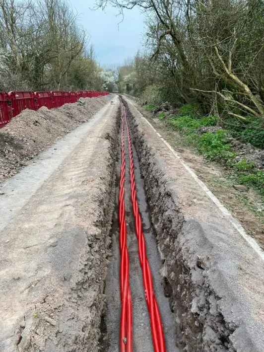 Red utility cables installed in a long trench dug along a rural roadside with trees and red safety barriers