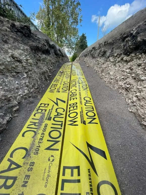 Yellow caution tape warning "Electric Cable Below" laid in a narrow trench along a rural roadside under blue sky